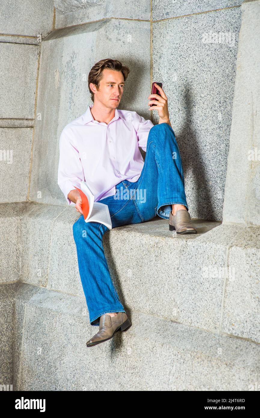 Detente a l exterieur. Portant une chemise a manches longues rose clair un Jean bleu des chaussures en cuir un jeune homme est assis tranquillement contre un mur de beton tenant Photo Stock