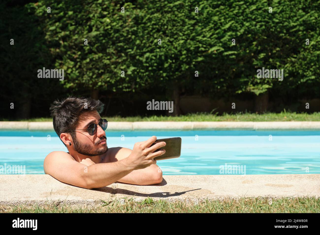 Jeune homme portant des lunettes de soleil, prenant un selfie dans une piscine. Banque D'Images