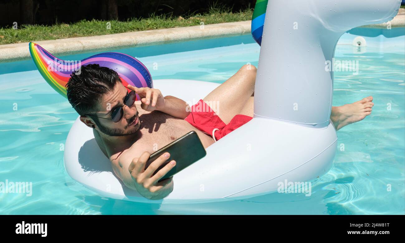 Jeune homme portant des lunettes de soleil prenant un selfie sur un anneau gonflable unicorn. Banque D'Images