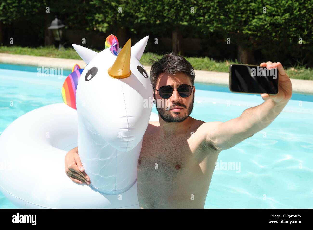 Jeune homme porte des lunettes de soleil prendre un selfie avec une licorne gonflable dans une piscine. Banque D'Images