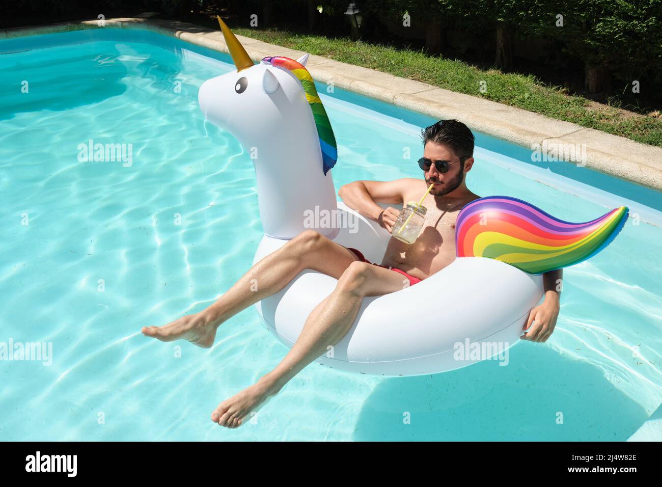 Un jeune homme avec des lunettes de soleil boit un rafraîchissement sur un anneau gonflable unicorn. Banque D'Images