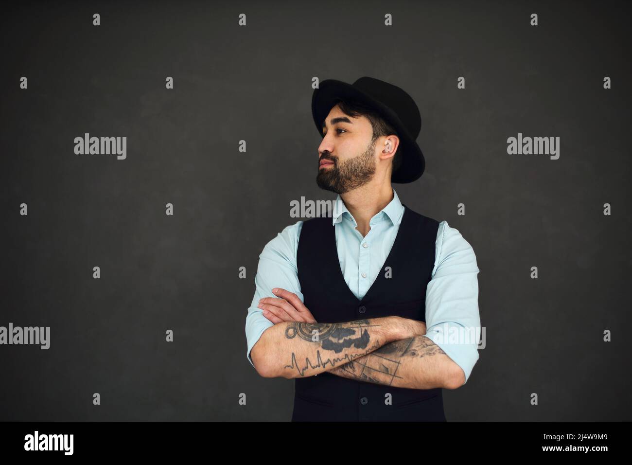 Portrait de studio de sexe masculin avec bras tatoués croisés, vêtu d'une chemise bleue et d'un chapeau noir. Jeune homme élégant sur fond sombre Banque D'Images