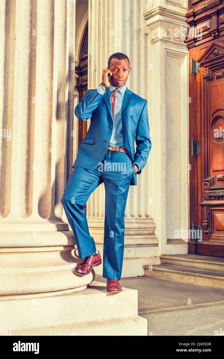 Robe de chambre bleue, cravate, chaussures en cuir, un jeune homme  d'affaires noir debout à l'extérieur de l'immeuble de bureau de style  vintage, appelant sur son téléphone mobile. INST Photo Stock -