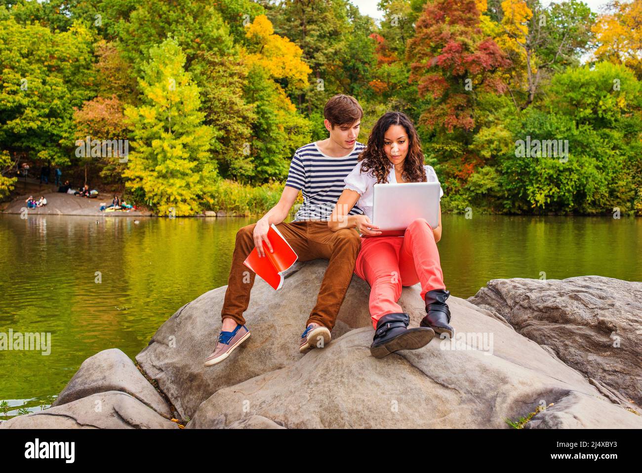 Jeune couple étudiant à l'extérieur près d'un lac, gars portant un t-shirt rayé, pantalon marron, baskets, tenant un livre, robe de fille en haut blanc, pantalon rouge, Banque D'Images