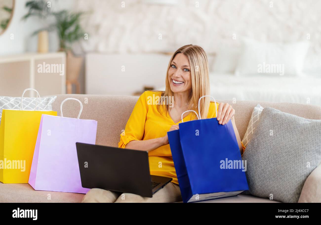 Une jeune femme blonde européenne satisfaite, vêtue de vêtements jaunes, avec un ordinateur portable et des looks dans un forfait shopping Banque D'Images