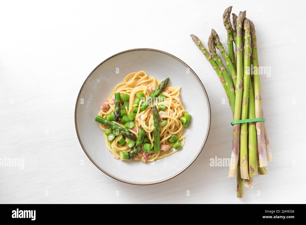 Asperges vertes avec spaghetti, bacon et sauce carbonara sur une assiette à côté d'un paquet de légumes crus sur une table en bois peinte en blanc, espace de copie, Banque D'Images