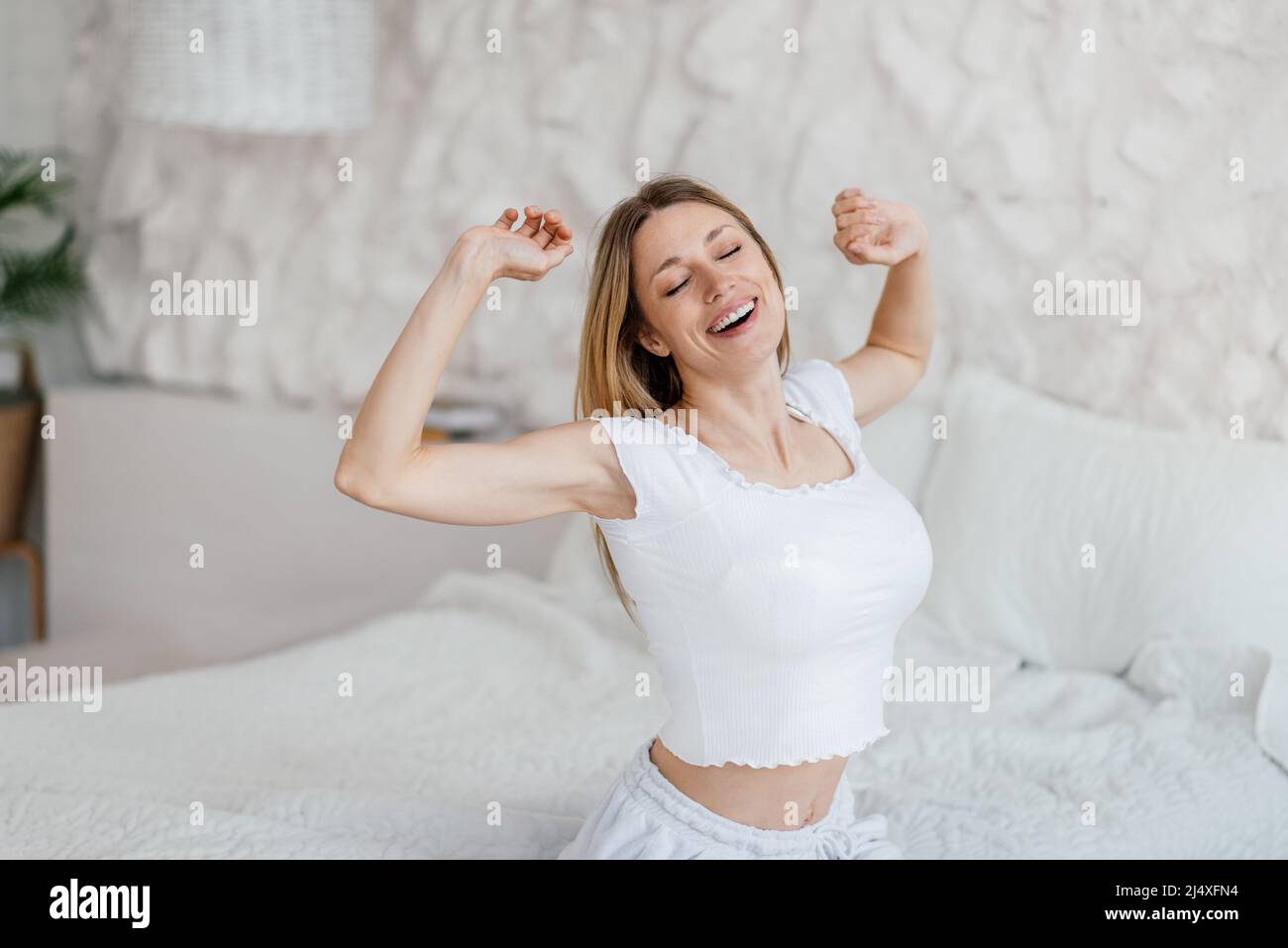 Gaie femme blonde européenne millénaire se réveille et étire le corps sur le lit seul dans l'intérieur de la chambre Banque D'Images