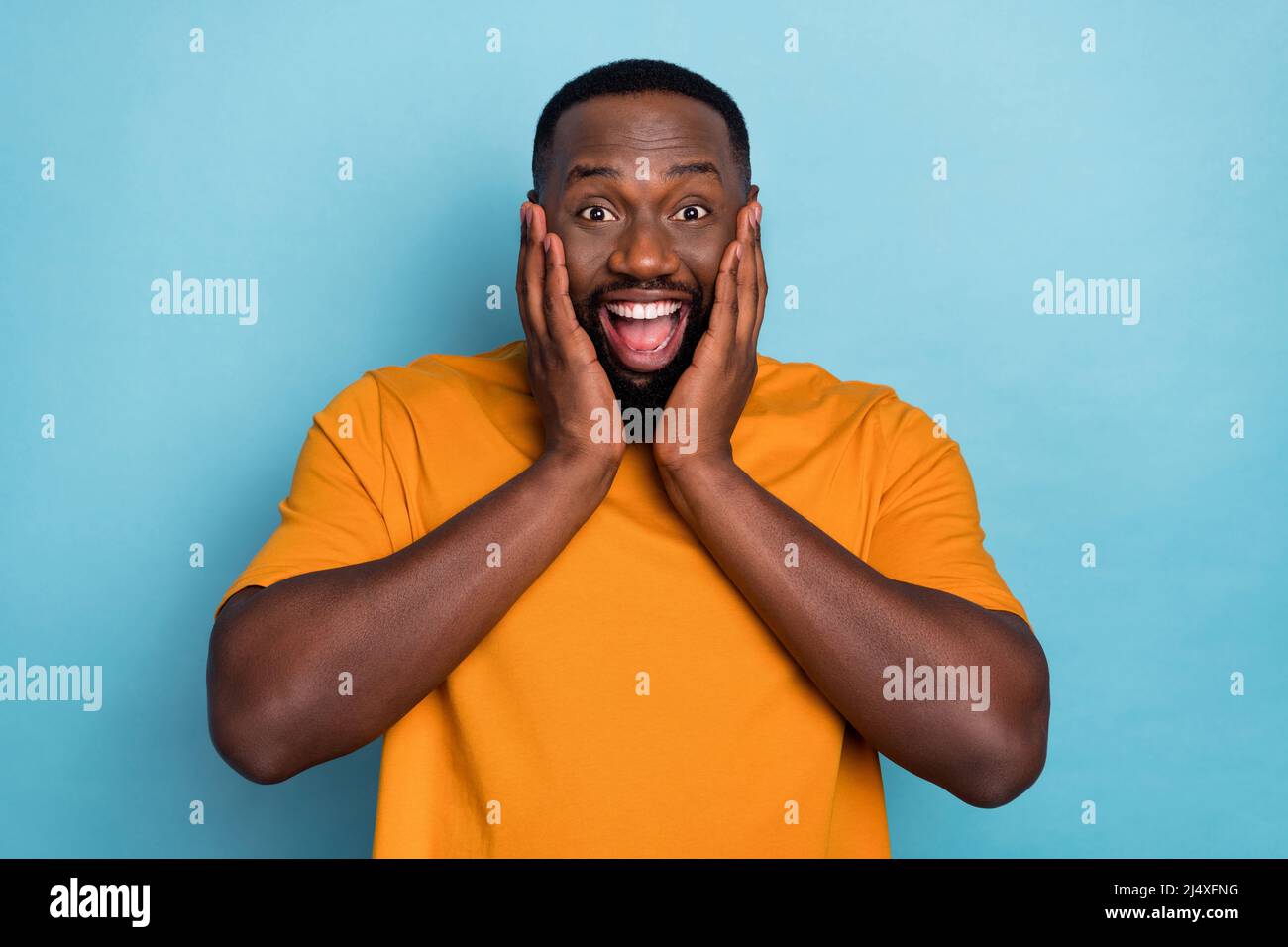 Photo de funky choqué gars habillé t-shirt orange souriant bras joues isolé couleur bleu fond Banque D'Images