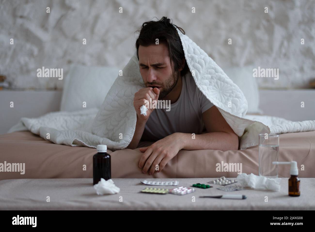Triste homme européen millénaire fatigué avec le chaume se trouve sous les couvertures, la toux et traité dans l'intérieur de la chambre avec des pilules Banque D'Images