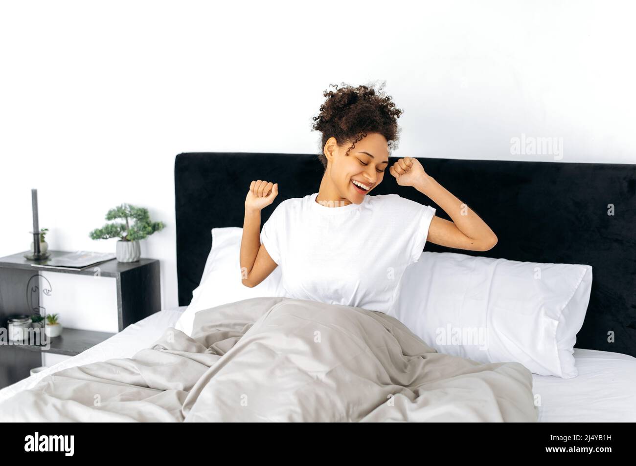 Réveil du matin. Bonne jolie fille afro-américaine à poil dur dans un t-shirt blanc, se réveille, se sourit les mains, commence une nouvelle journée avec une grande humeur, sourit, se couche au lit Banque D'Images