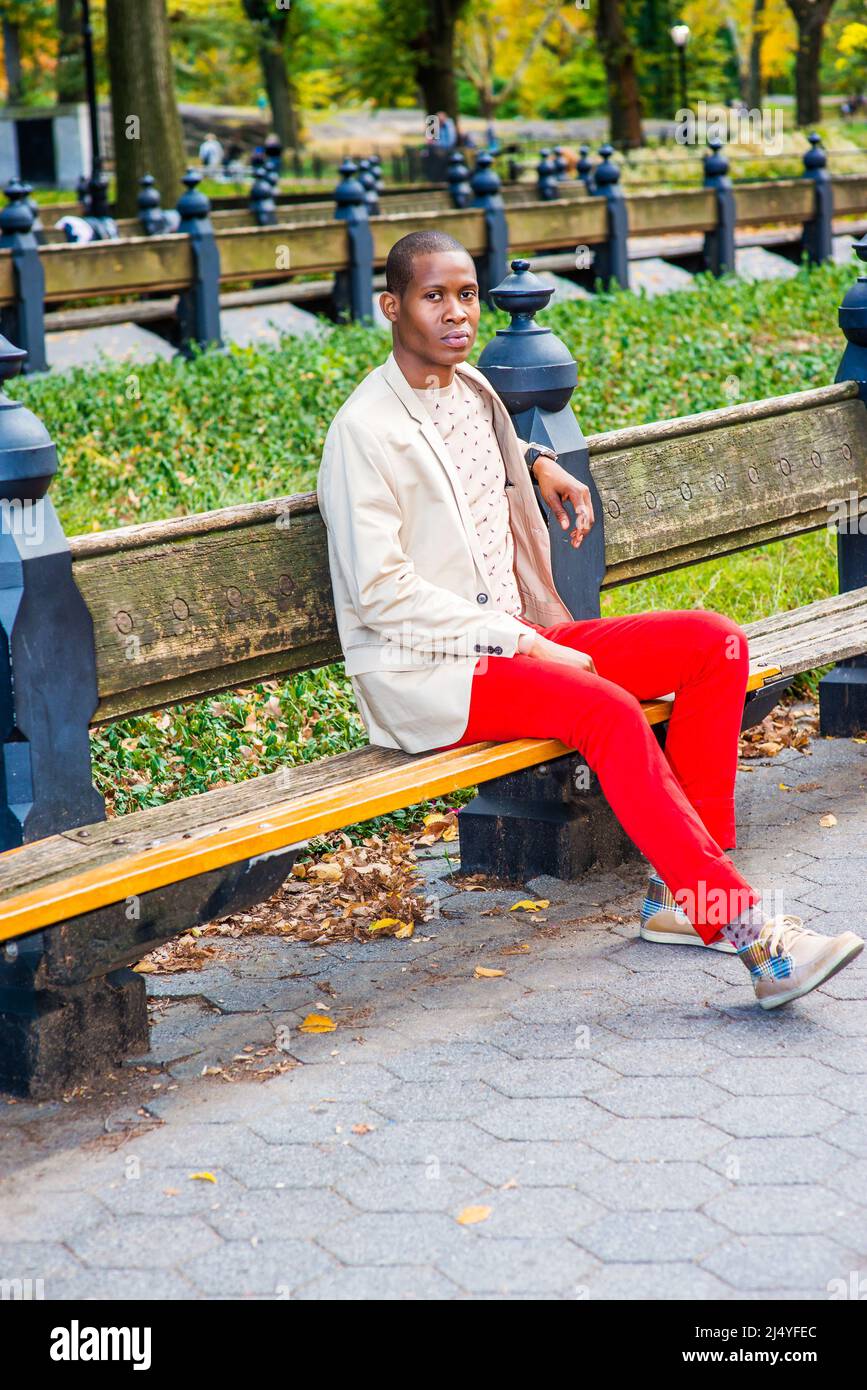 Homme qui vous attend. Déguisements en blazer crème, chandail sans collerre, pantalon rouge, baskets, un jeune homme noir est assis sur le banc sur le parc, lookin Banque D'Images