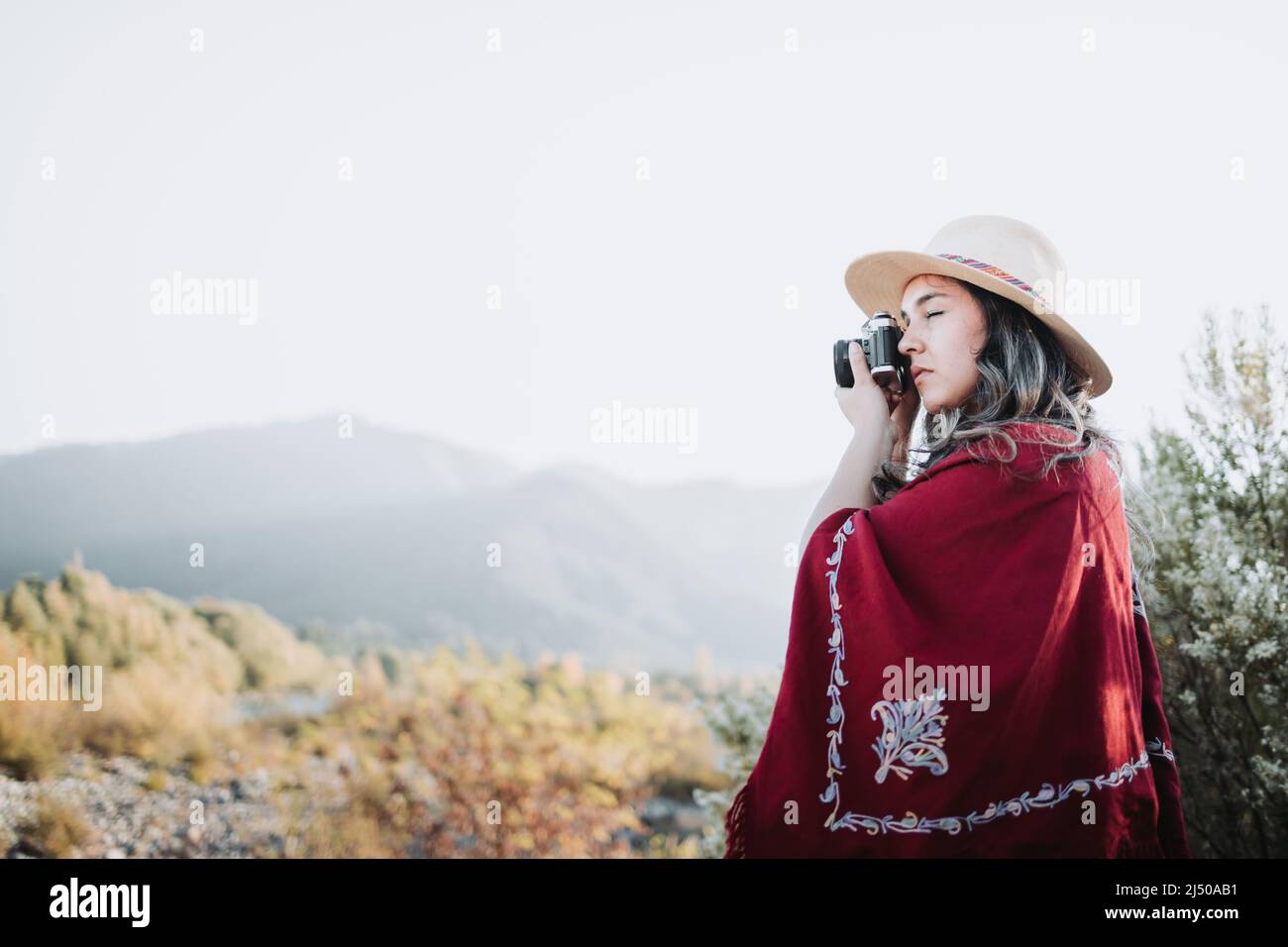 Été jeune femme portant un poncho rouge et un chapeau, utilisant un appareil photo vintage pour prendre des photos dans un espace naturel. Banque D'Images