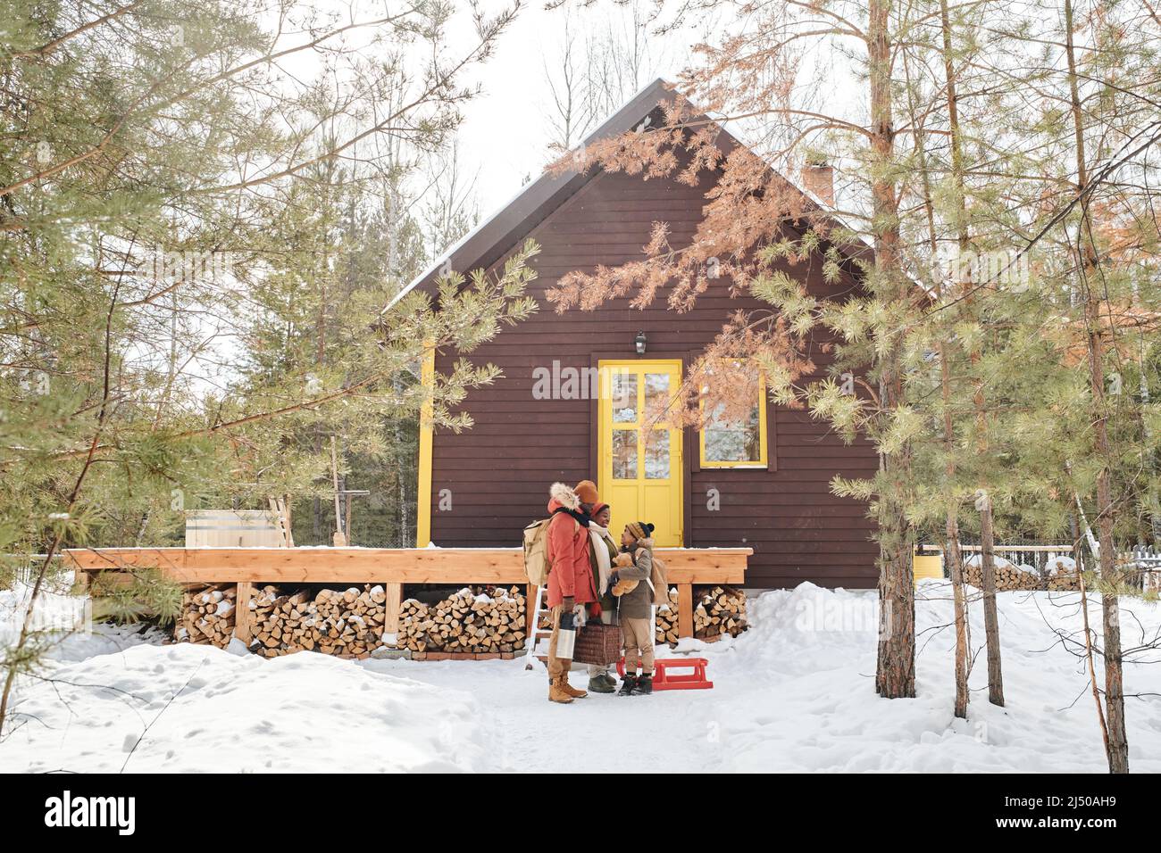 Famille contemporaine de parents et petit fils en vêtements d'hiver debout près de leur maison de campagne située dans les bois profonds parmi les pinetrees Banque D'Images