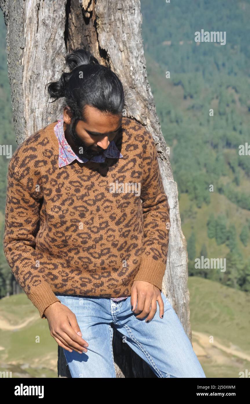 Un jeune homme élégant avec un petit pain à cheveux en regardant vers le bas tout en posant avec appuyé contre l'arbre Banque D'Images