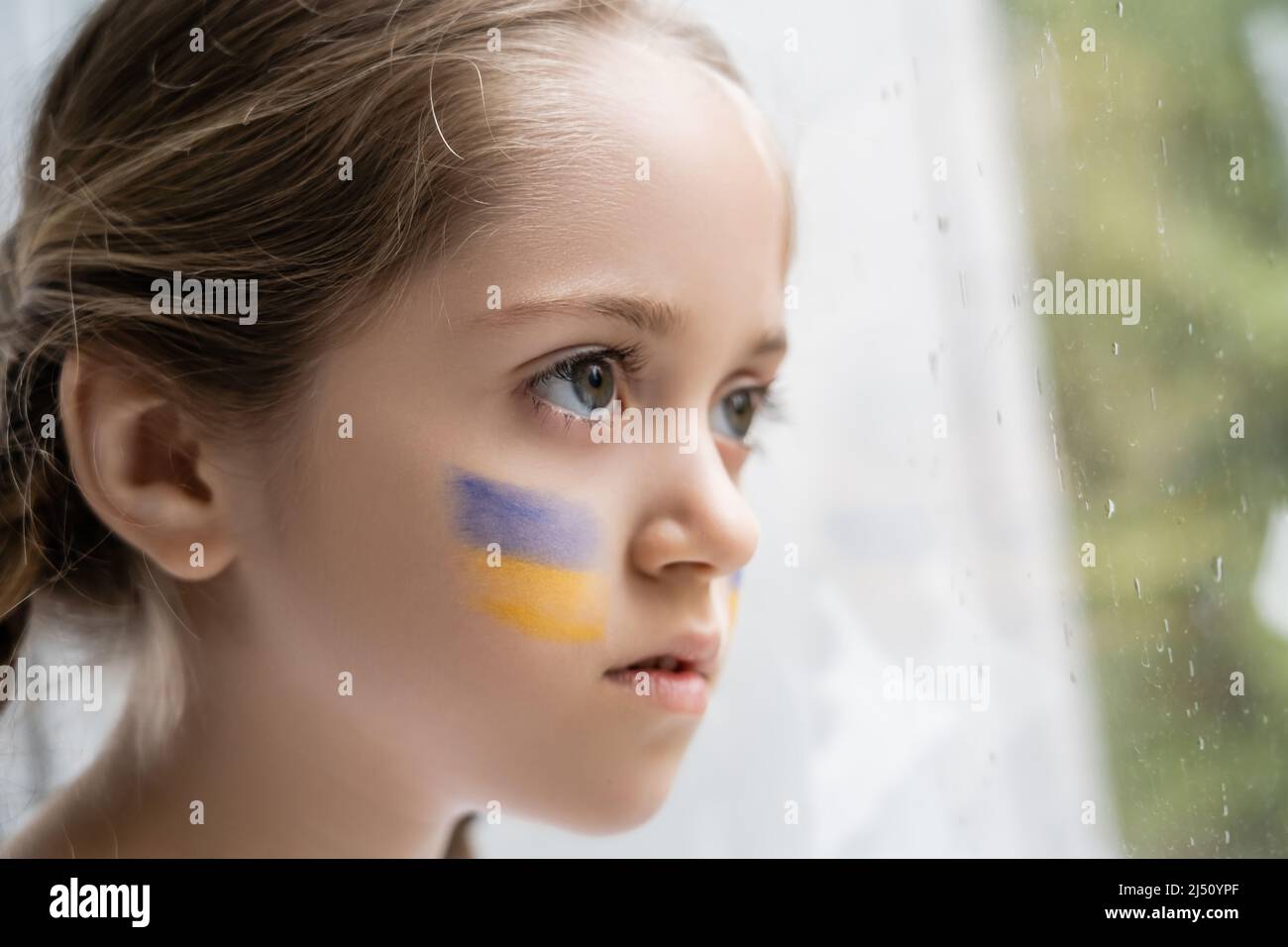 portrait de fille avec drapeau ukrainien sur le visage regardant loin par la fenêtre avec des gouttes de pluie Banque D'Images