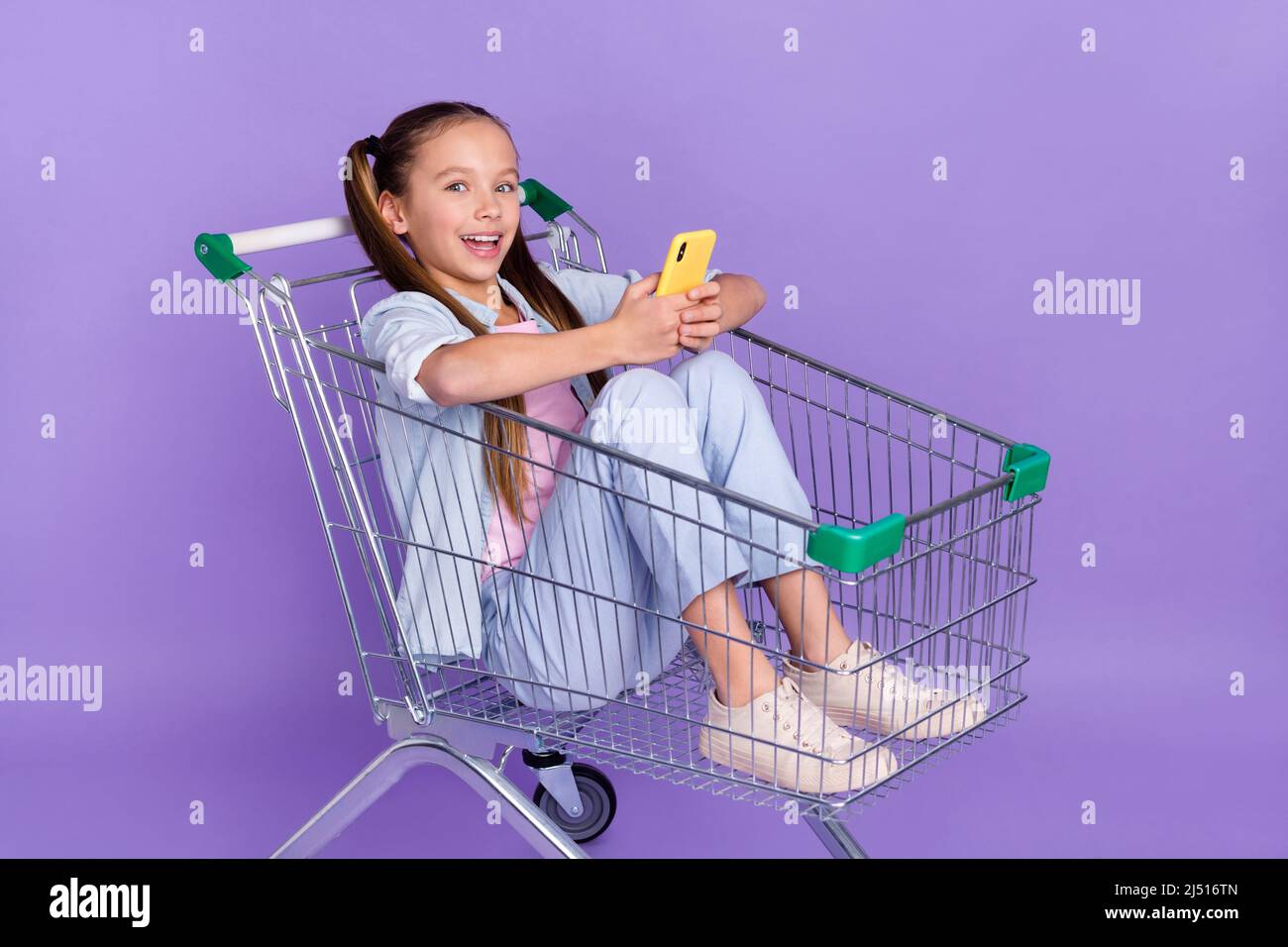 Photo de la petite écolière très impressionnée habillé jeans chemise conduite plateau shopping dactylographie gadget isolé violet couleur fond Banque D'Images