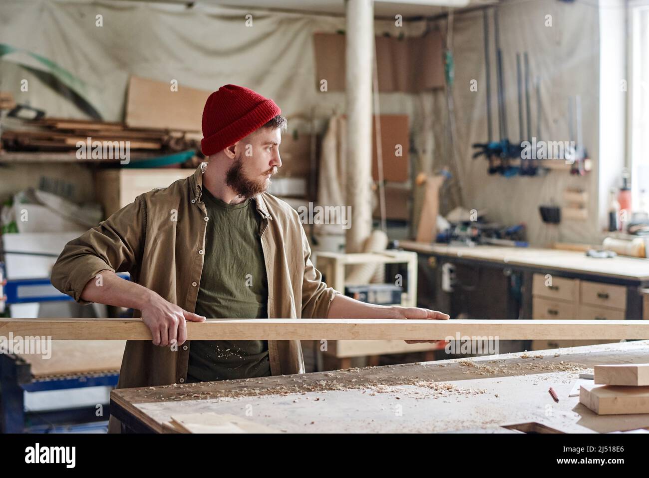 Jeune charpentier caucasien avec la barbe sur le visage portant la casquette tricotée rouge prenant la planche de bois pour couper et polir Banque D'Images