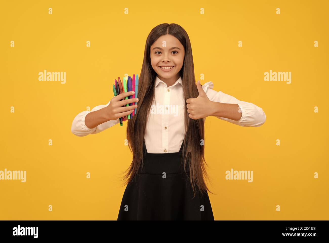 Happy girl enfant tenir les stylos feutre pointe montrant les pouces vers le haut mouvement de la main fond jaune, satisfait Banque D'Images