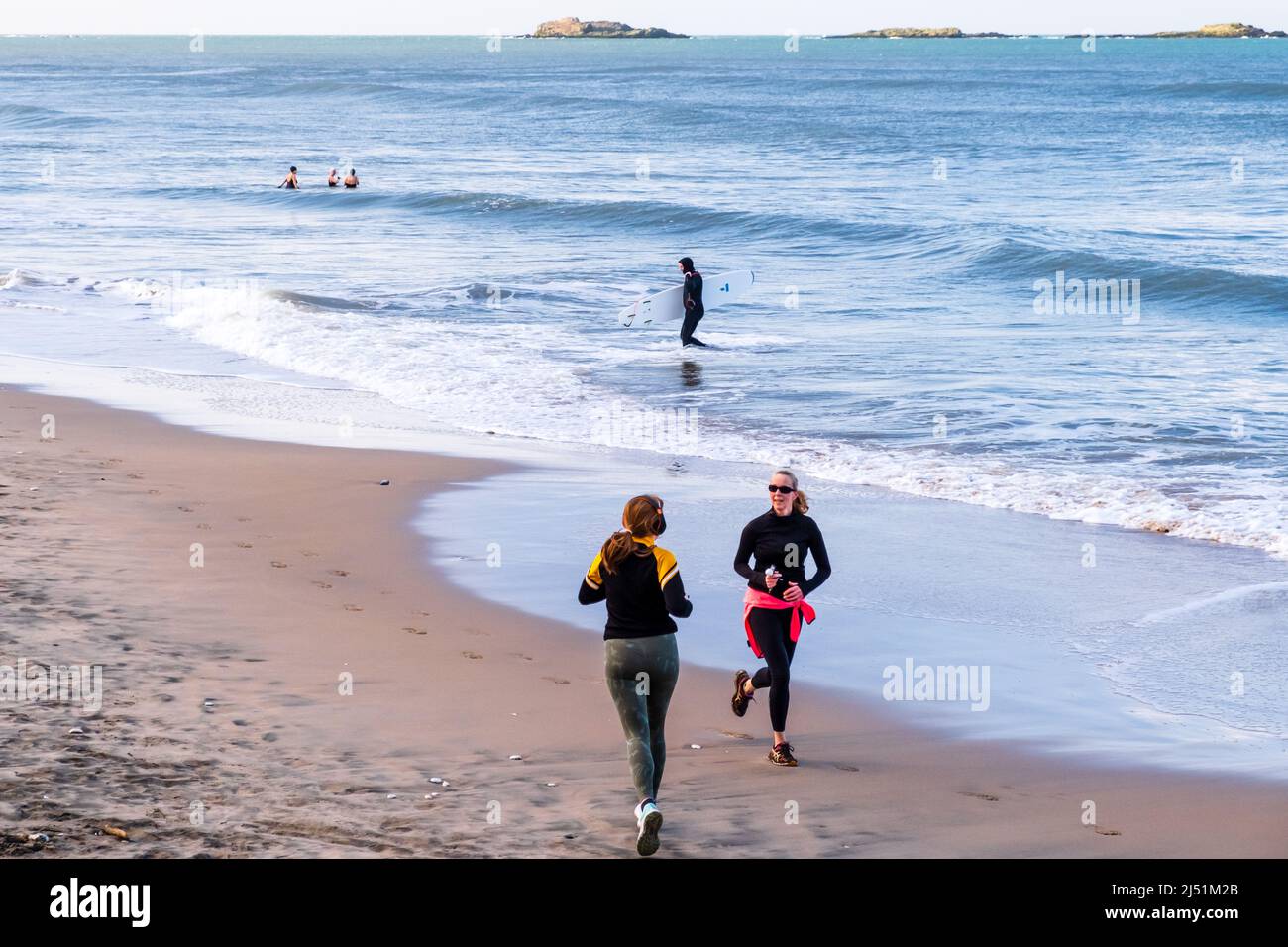 Deux coureurs se passant l'un l'autre, un surfeur avec sa planche quittant la mer et trois dames nageant à la plage de White Rocks, Portrush, County Antrim. Banque D'Images