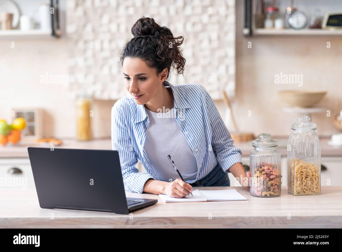 Latino femme écrivant une nouvelle recette regardant des leçons en ligne sur ordinateur portable dans la cuisine lumineuse intérieur, espace de copie Banque D'Images