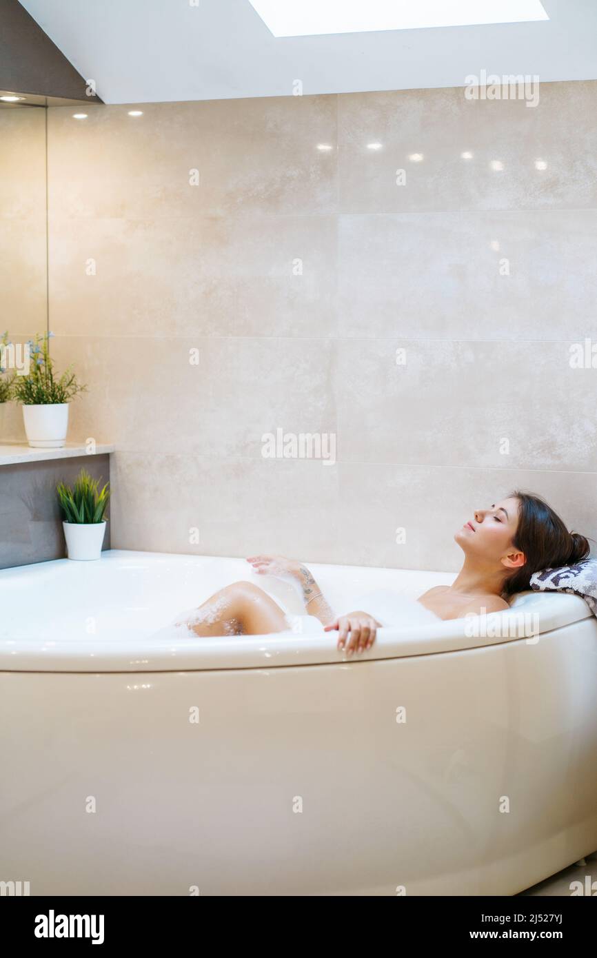 Jeune femme nue couché sur une serviette dans la baignoire à bulles blanches avec les yeux fermés et profiter. Vue sur le côté portrait d'une femme agréable qui se repose dans la salle de bains. Concept o Banque D'Images