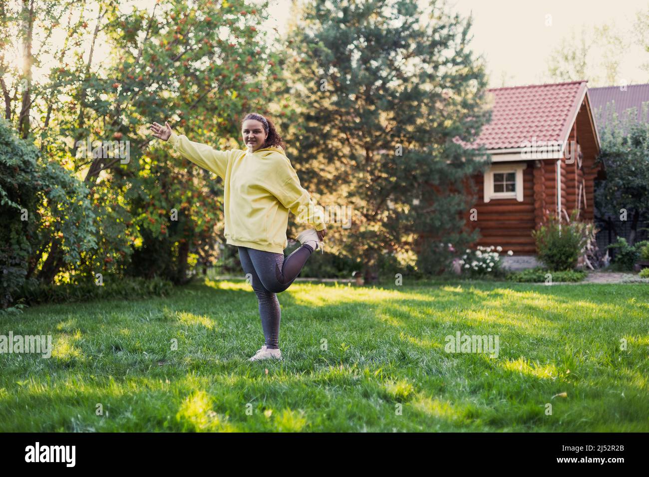 Femme sur l'herbe fraîche s'étirant porter des vêtements de sport avec un pied et la main sur l'arrière-cour pleine de lumière du soleil avec maison de campagne et haut Banque D'Images