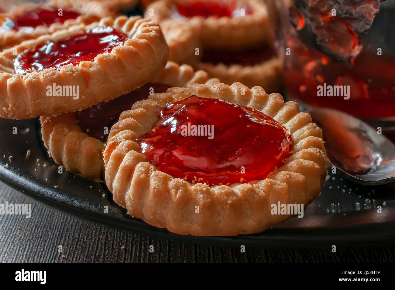 tartelez les biscuits avec de la confiture de fraise sur une assiette noire avec une cuillère et un verre avec de la confiture sur le côté, vue latérale macro-photographie de nourriture Banque D'Images