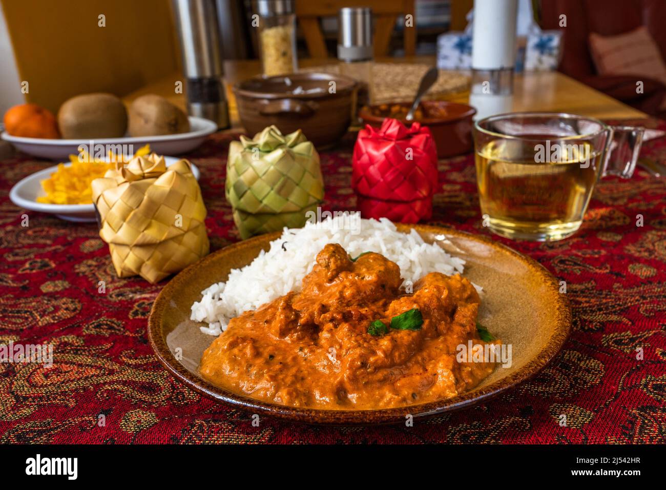 Tikka masala de poulet indien épicé avec riz sur l'assiette, boîte à 3 épices en bambou, tasse de thé, fruit sur nappe rouge sur la table. Banque D'Images