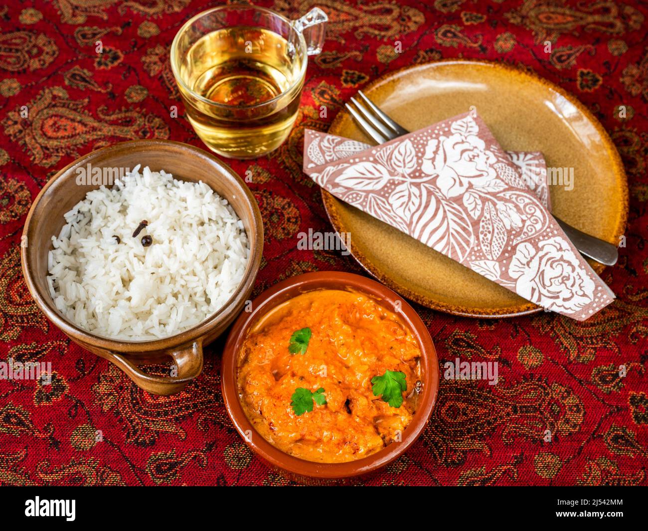 Tikka masala de poulet épicé avec riz cuit dans un bol de 2, tasse de thé, assiette avec serviette et fourchette sur nappe rouge. Cuisine indienne. Banque D'Images