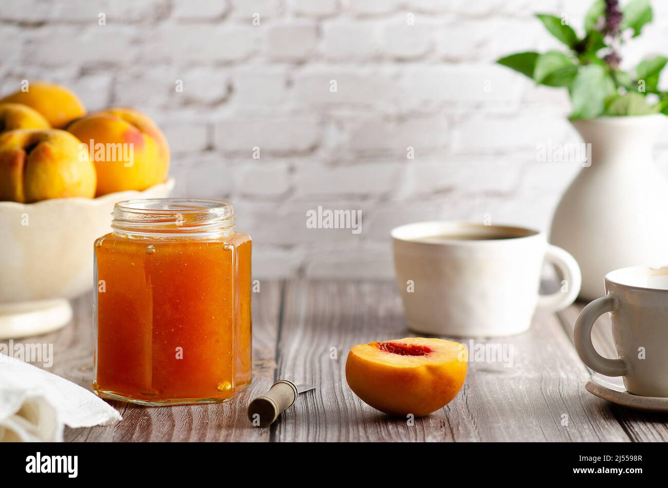 Confiture de pêches maison dans un bocal avec des pêches fraîches, deux tasses de café et un vase avec des feuilles vertes, sur un fond de table en bois. Banque D'Images