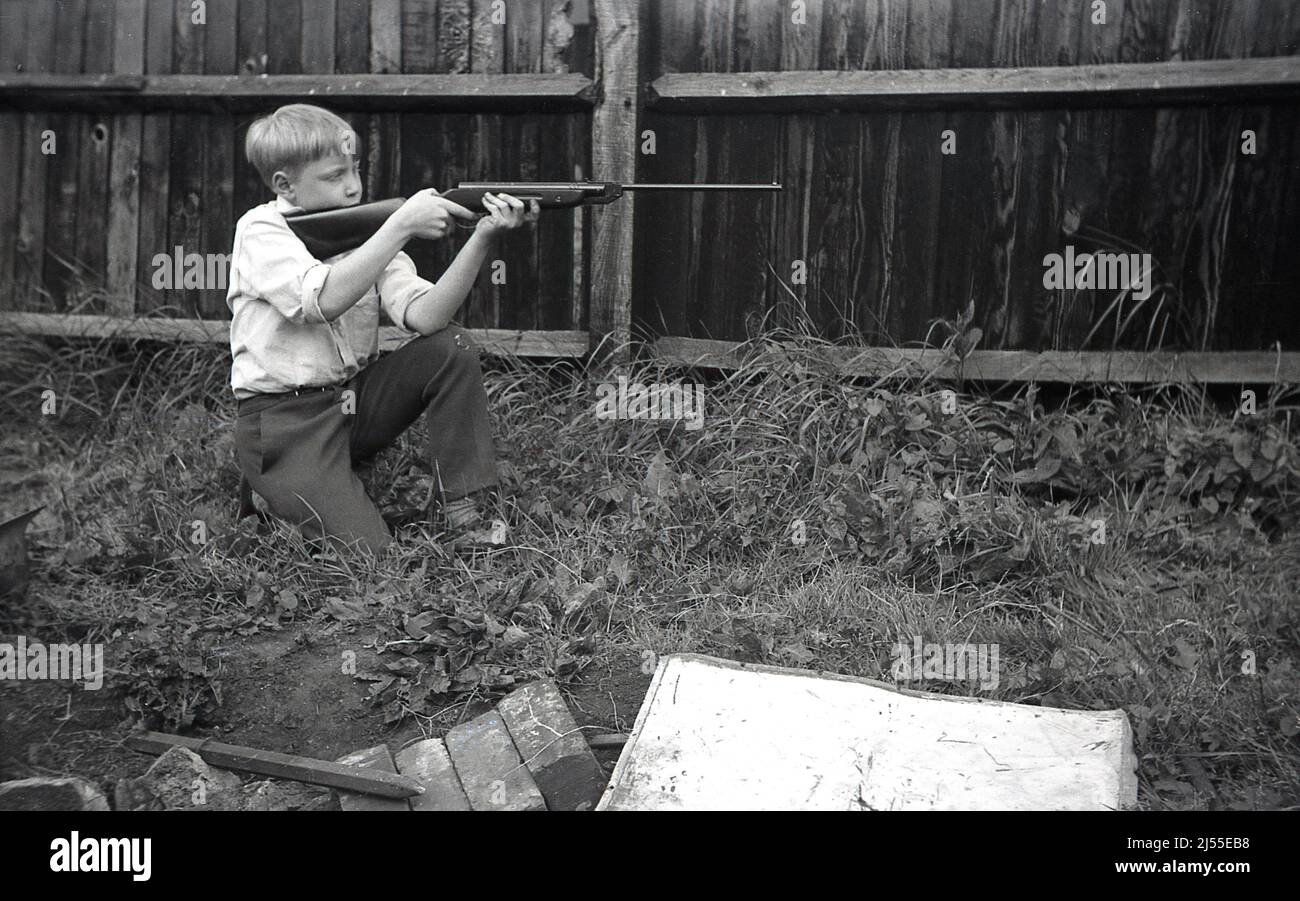 1960s, historique, à l'extérieur dans un jardin à l'arrière, à côté d'une clôture en bois, un garçon s'agenouillant, tenant une carabine à air comprimé dans les deux mains dans une position de tir ou de tir, peut-être une Diana modèle 27, Angleterre, Royaume-Uni. Banque D'Images