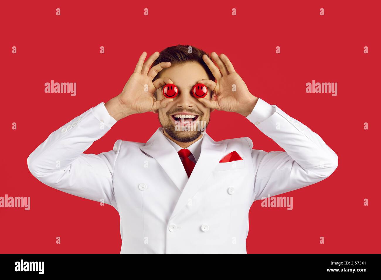 Beau homme chanceux souriant et couvrant ses deux yeux avec deux jetons rouges de poker en mains, debout sur fond rouge studio, copyspace . Casino et Banque D'Images