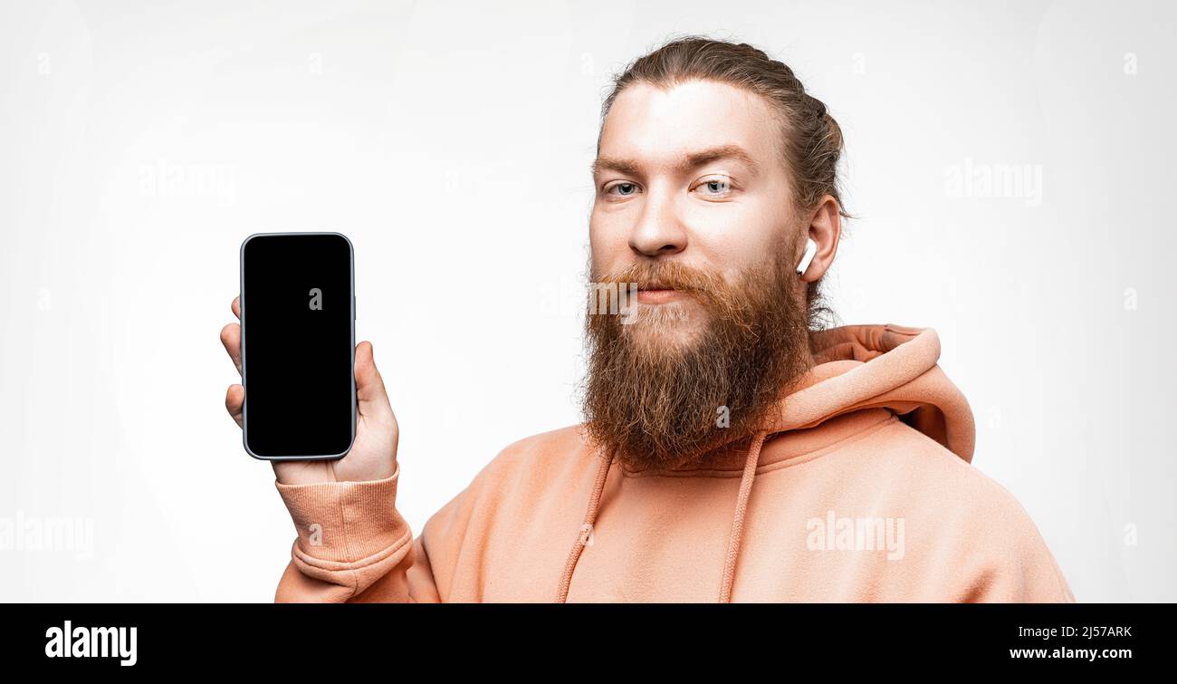 Scandinave beau homme sérieux tenant un téléphone avec un écran noir maquette avec casque sans fil isolé sur fond gris. Homme calme avec la ginge Banque D'Images