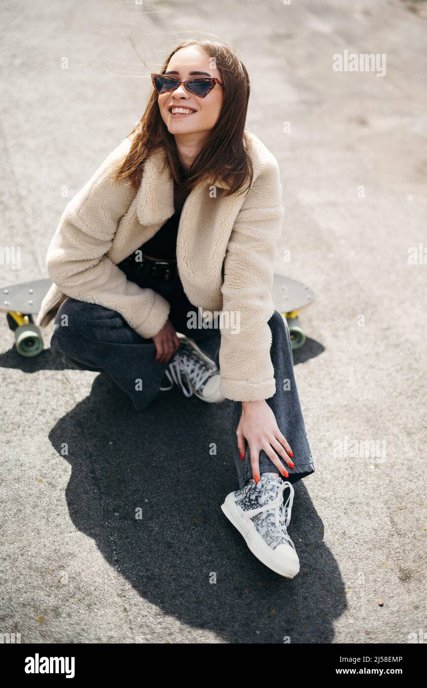 Joyeuse belle femme dans des vêtements tendance s'amuser tout en étant  assis sur le skateboard à l'extérieur. Zone urbaine avec bâtiment moderne  sur fond. Modes de vie Photo Stock - Alamy