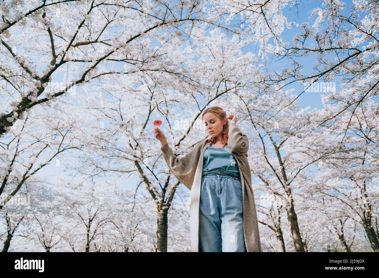 Jeune femme profitant de la nature au printemps. Pique-nique avec un verre de vin sous les cerisiers en fleurs. La femme aime la vie et un beau parc avec des cerisiers Banque D'Images