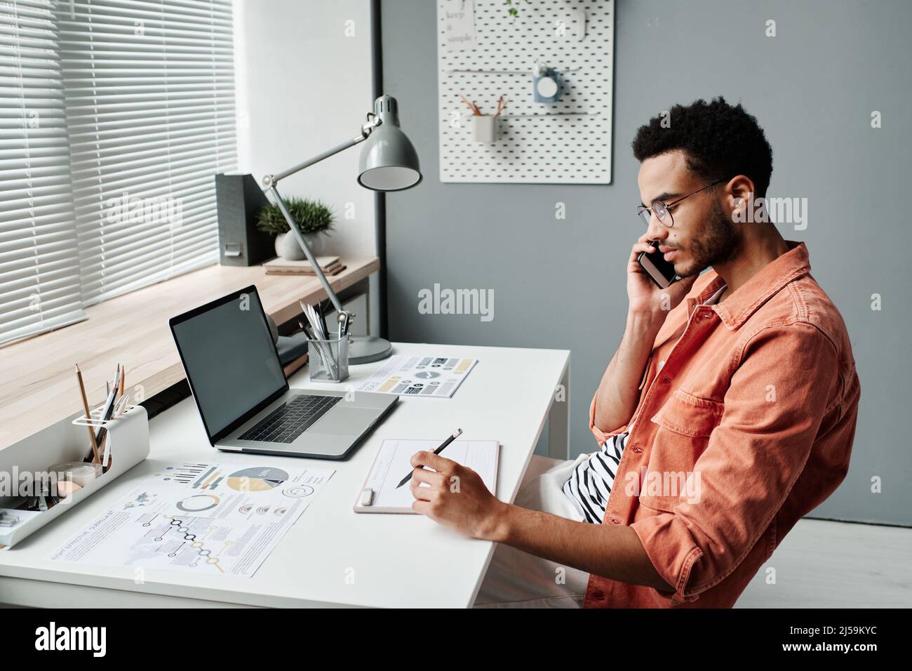 Jeune homme de course mixte sérieux avec barbe de 3 jours assis au bureau avec un ordinateur portable et des papiers et planification de la réunion tout en parlant au client par téléphone Banque D'Images
