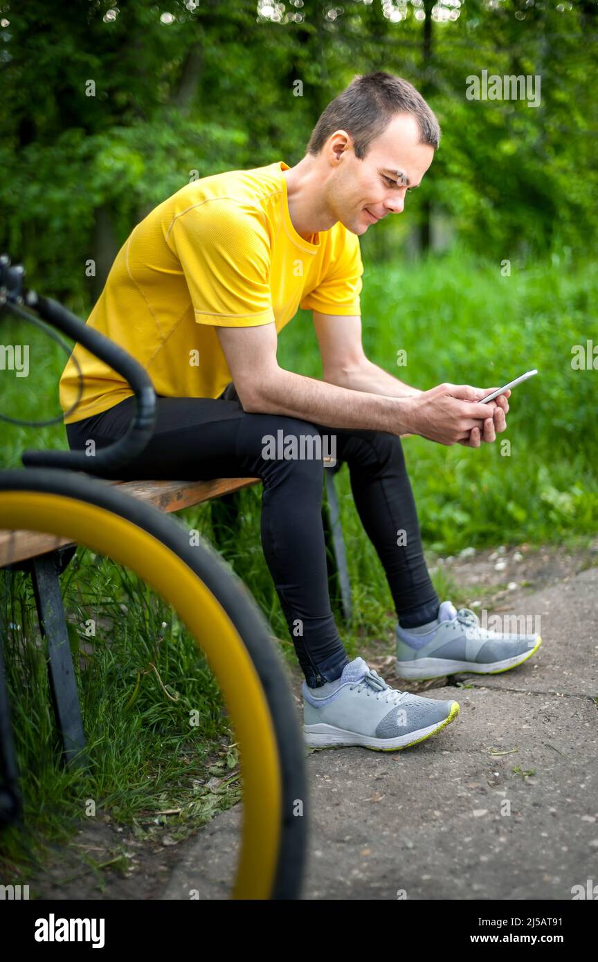 Un jeune homme assis sur un banc dans un parc public communique par communication mobile. En arrière-plan avec son vélo. Parmi les arbres et la végétation. Banque D'Images