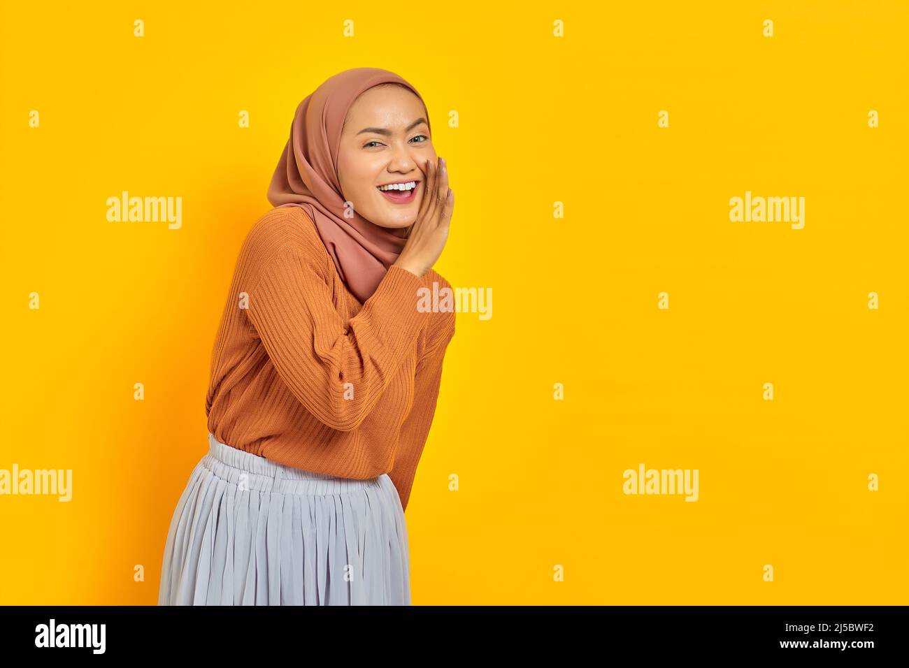 Belle jeune femme asiatique en chandail brun et hijab main sur la bouche en racontant le secret, en chuchotant des potins, en regardant la caméra avec l'expression joyeuse iso Banque D'Images