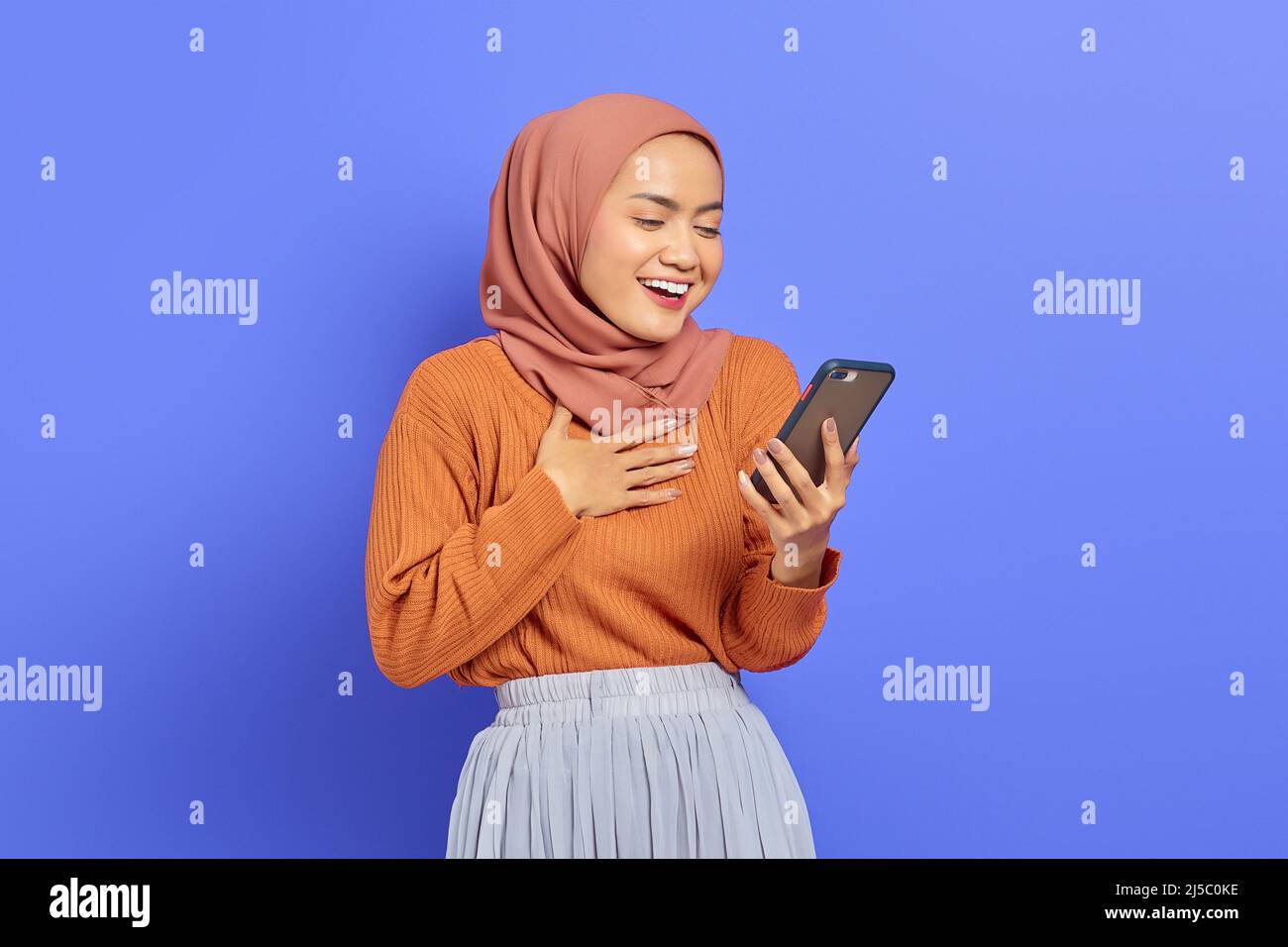 Portrait d'une femme asiatique souriante en chandail brun et hijab tenant sur un téléphone portable avec la main sur la poitrine isolée sur fond violet. Péopl Banque D'Images