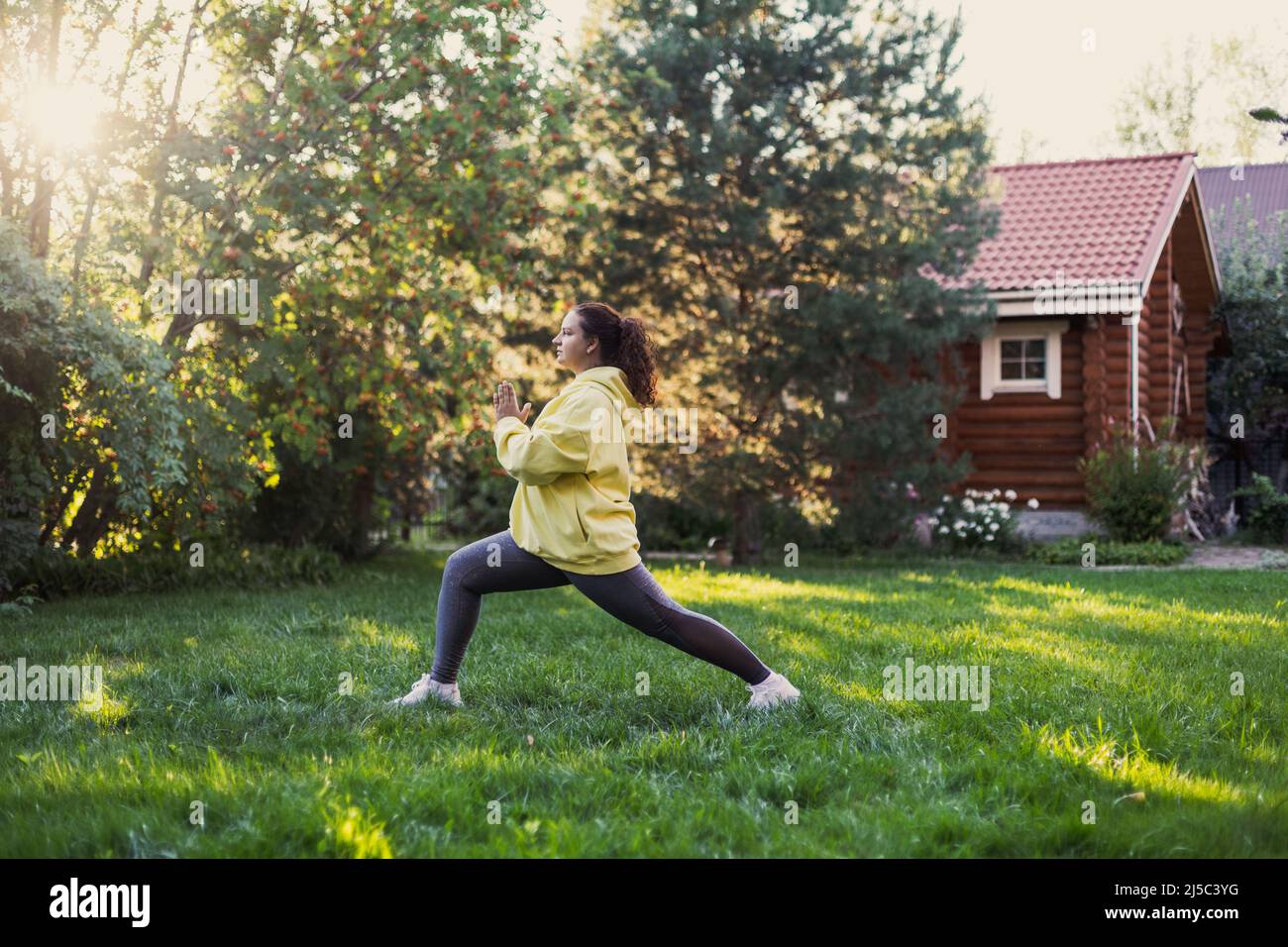 Femme grasse d'âge moyen faisant des exercices de yoga dans les vêtements de sport regardant loin sur l'herbe fraîche sur l'arrière-cour avec maison de campagne en bois et de grands arbres dedans Banque D'Images
