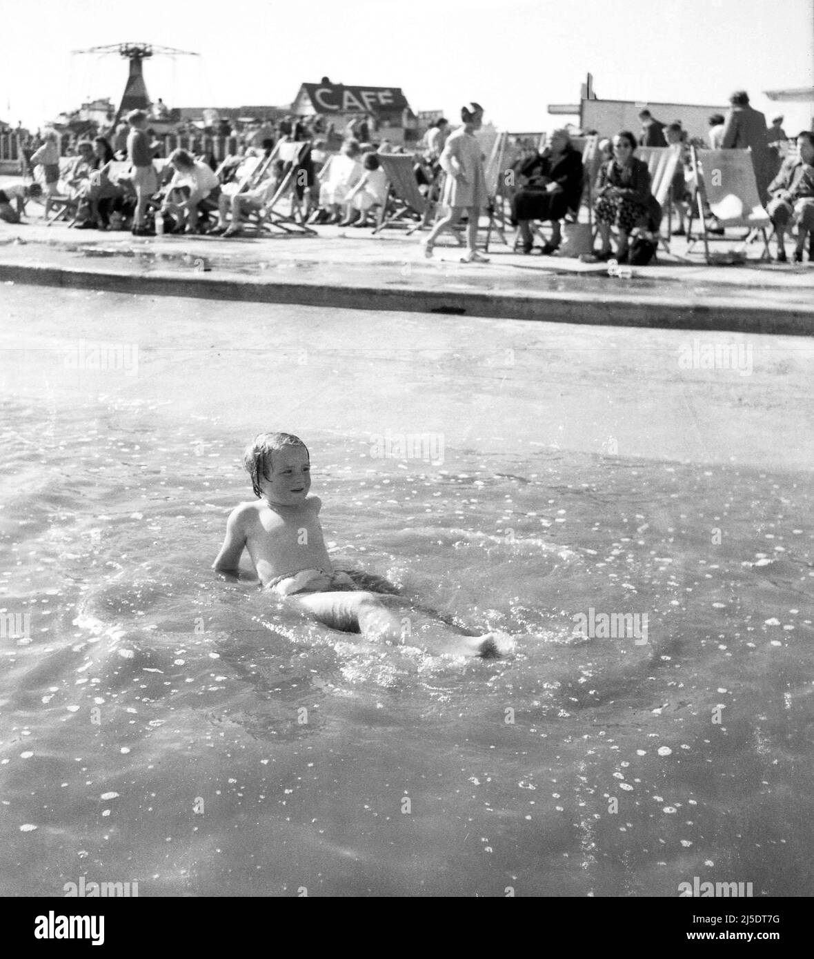 1960, historique, une petite fille couchée dans des eaux peu profondes à un Lido extérieur, avec des personnes assises dans des chaises longues sur le côté, Angleterre, Royaume-Uni. Banque D'Images