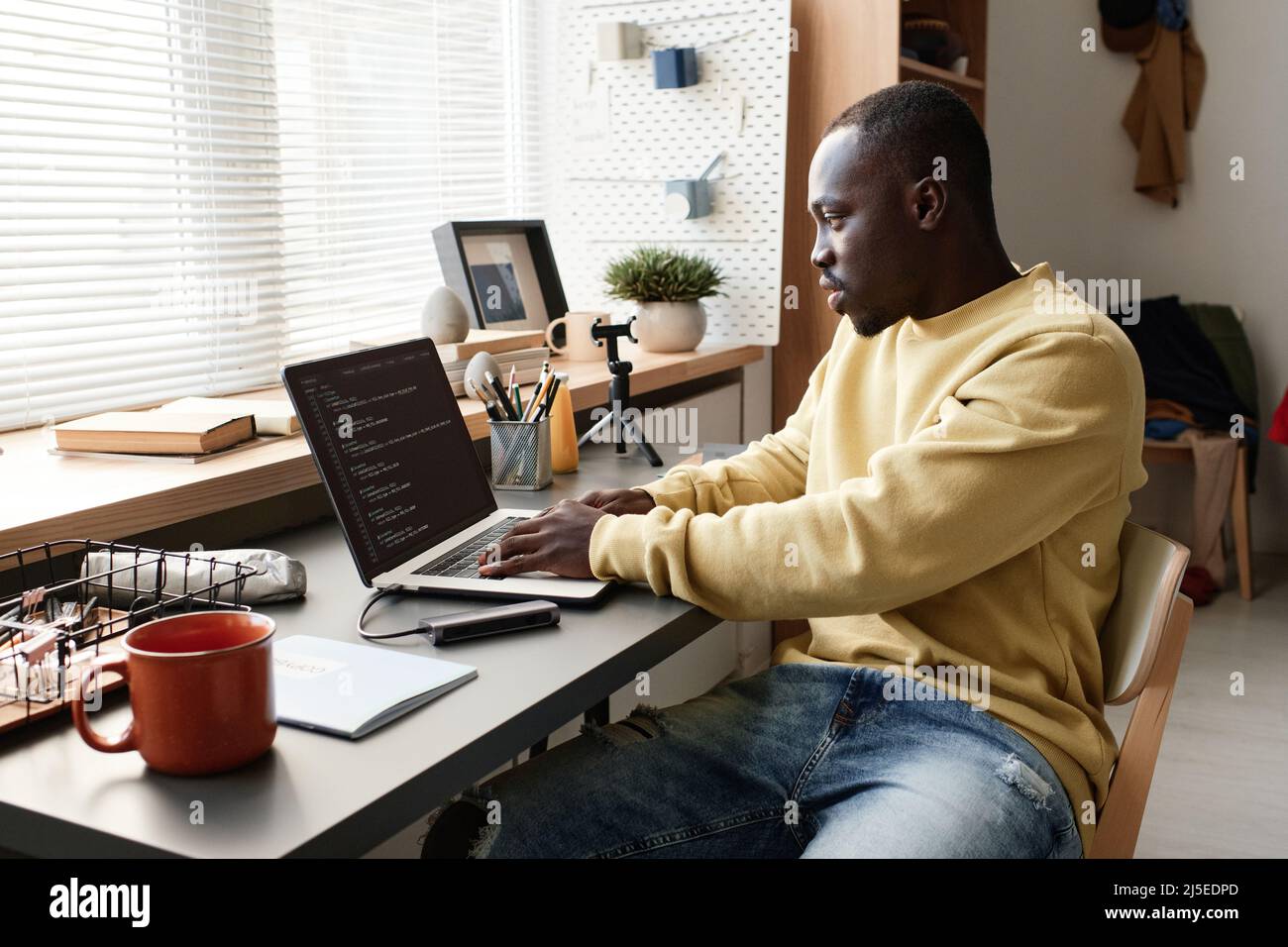 Sérieux youn Noir étudiant gars en chandail jaune assis au bureau et codage Java script sur ordinateur portable Banque D'Images