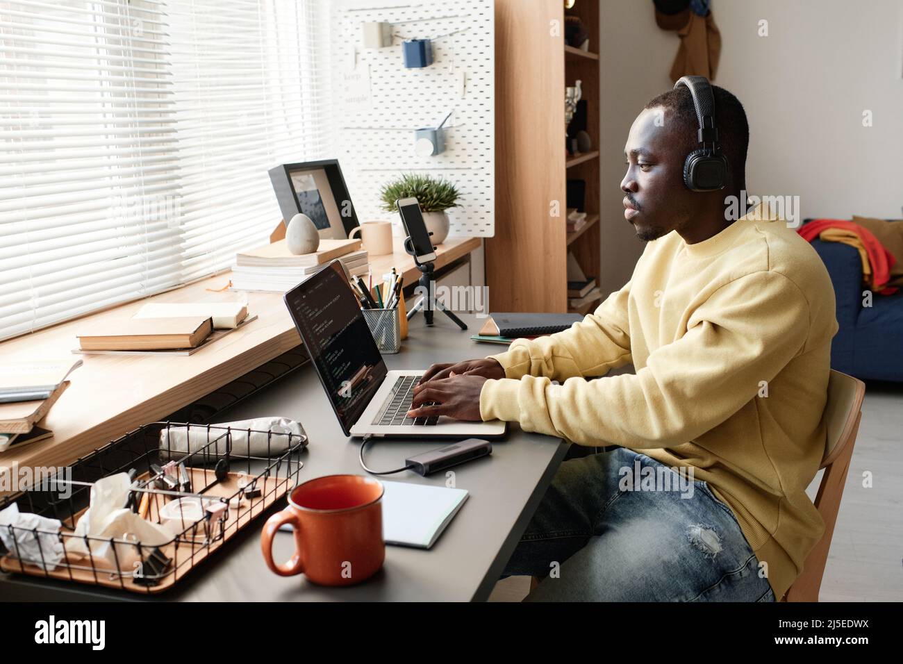 Jeune spécialiste afro-américain du codage concentré dans les écouteurs assis à table contre la fenêtre et utilisant un ordinateur portable lors de la création d'une application Banque D'Images