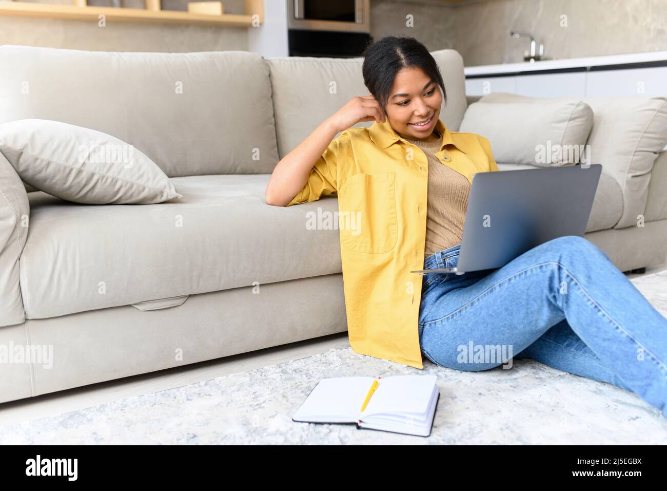 Charmante et calme, cette femme afro-américaine est assise sur le sol dans un salon confortable avec un ordinateur portable. Elle aime passer un week-end paresseux dans une atmosphère détendue, travailler à distance à la maison, discuter ou étudier en ligne Banque D'Images