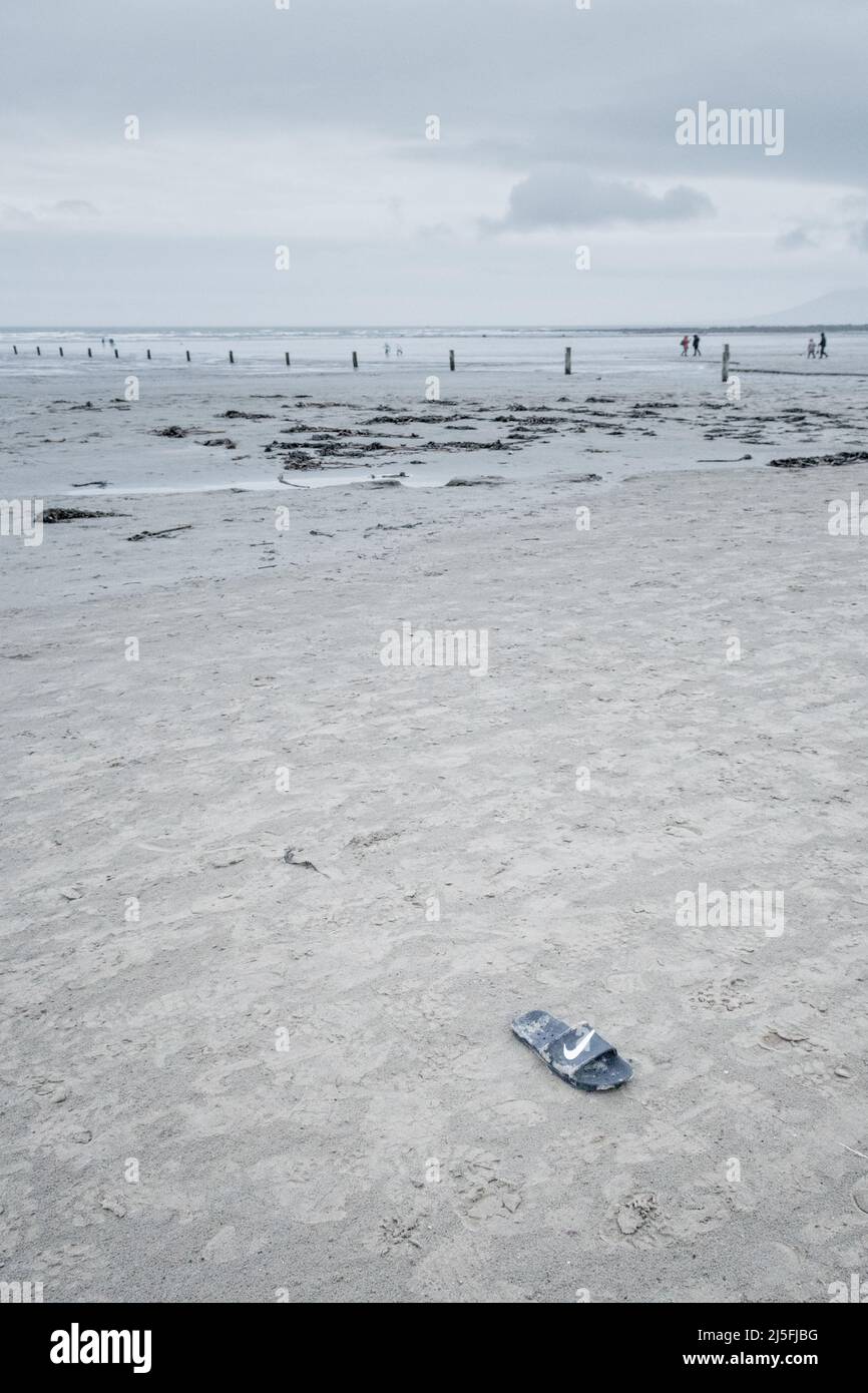 Un seul santal Nike délavé sur la plage de Tirella, en Irlande du Nord ; personnages éloignés en arrière-plan ; après-midi d'hiver couvert par le froid ; couleurs pâles. Banque D'Images