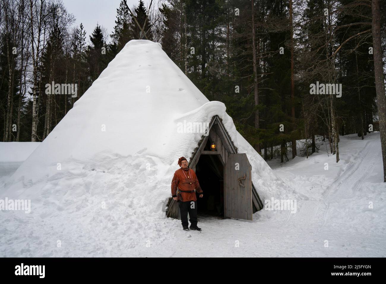 Rovaniemi, Finlande - le 18th mars 2022 : un homme sami en vêtements traditionnels, debout à la porte de sa maison traditionnelle en bois sâme enneigée, appelle Banque D'Images