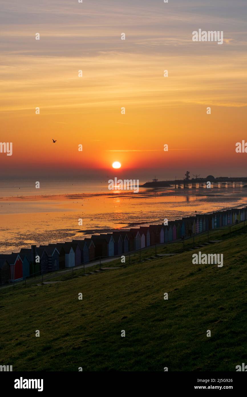 HERNE BAY : le soleil du matin qui se lève sur la mer le dimanche de Pâques avec des cabanes de plage vues en premier plan et la jetée en arrière-plan. Banque D'Images