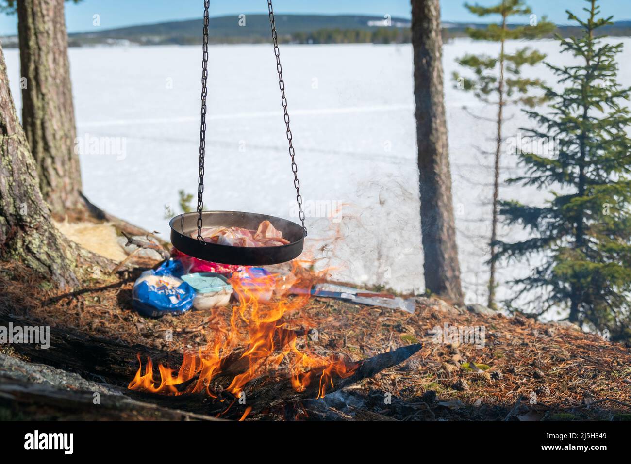 Poêle à frire pleine de bacon suspendu sur un feu de camp, lac gelé et ciel bleu en arrière-plan. Photo de Vasternorrland Suède. Mise au point sur le premier plan. Banque D'Images