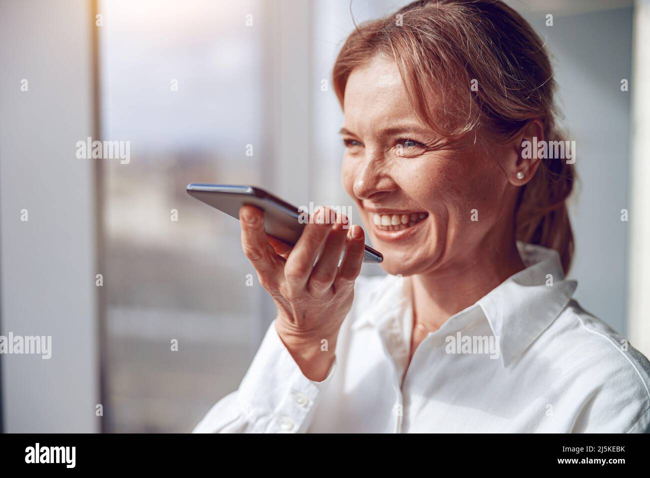 une femme mûre et souriante utilise un smartphone pour enregistrer un massage vocal à son partenaire d'affaires Banque D'Images