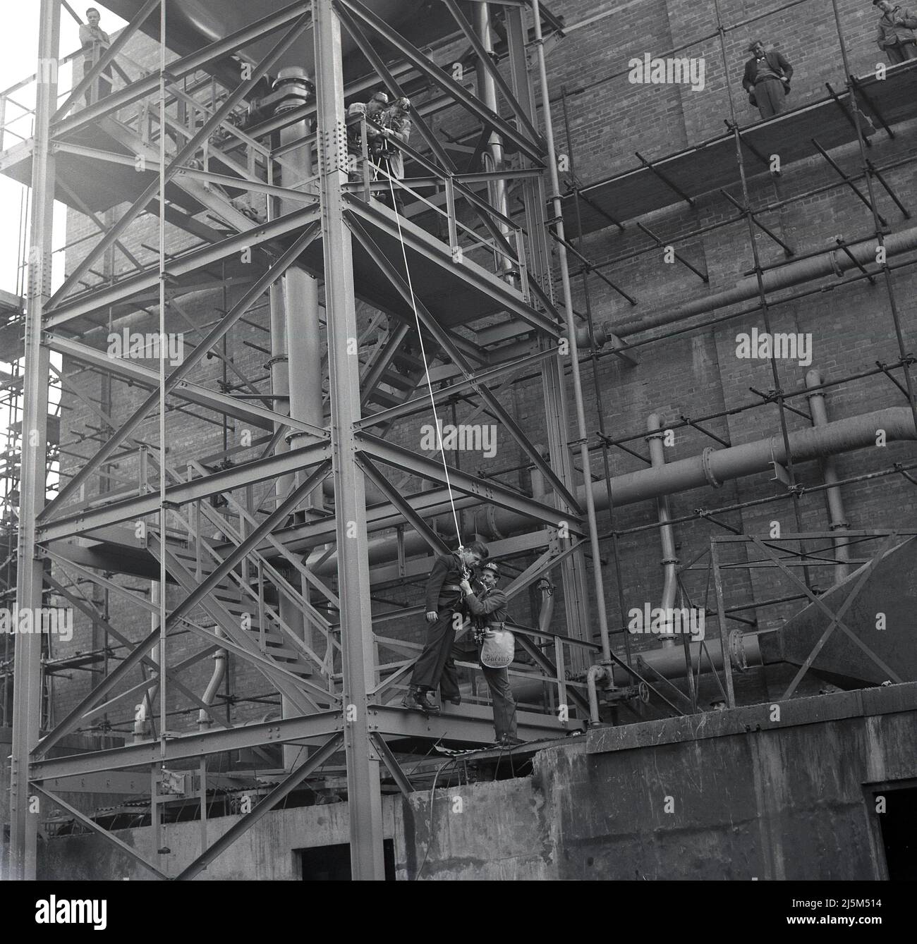 1950s, historique, forage de sécurité, ouvrier de l'acier étant abaissé à partir d'un palan de grande structure, étant tenu par un autre travailleur portant des appareils respiratoires, steelworks, Abbey Works, Margam, Port Talbot, Pays de Galles, Royaume-Uni. Banque D'Images